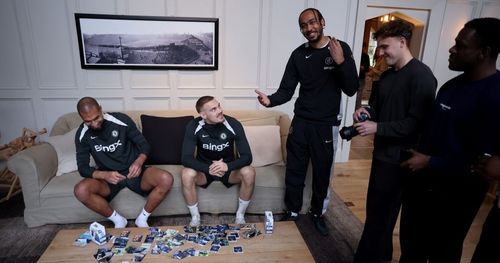 Five men in sportswear gathered in a living room with two sitting on a couch and the other three standing, among them a man holding a camera and trading cards spread on the coffee table.