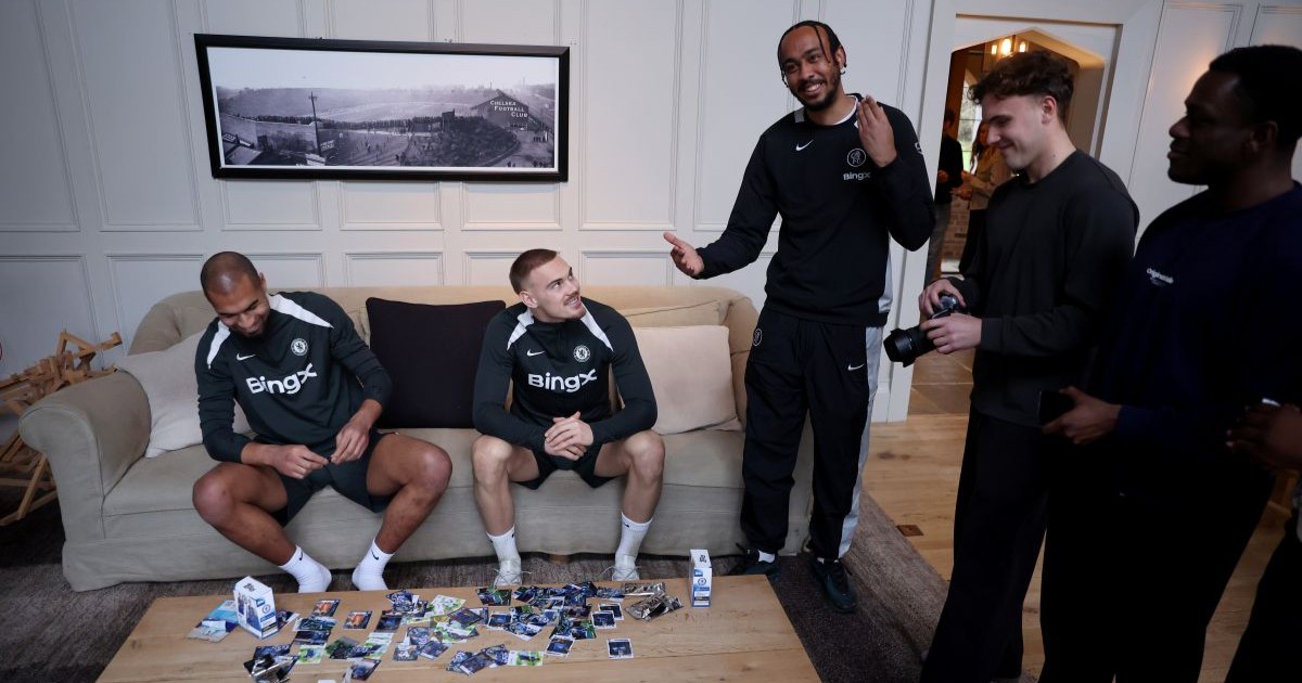 Five men in sportswear gathered in a living room with two sitting on a couch and the other three standing, among them a man holding a camera and trading cards spread on the coffee table.