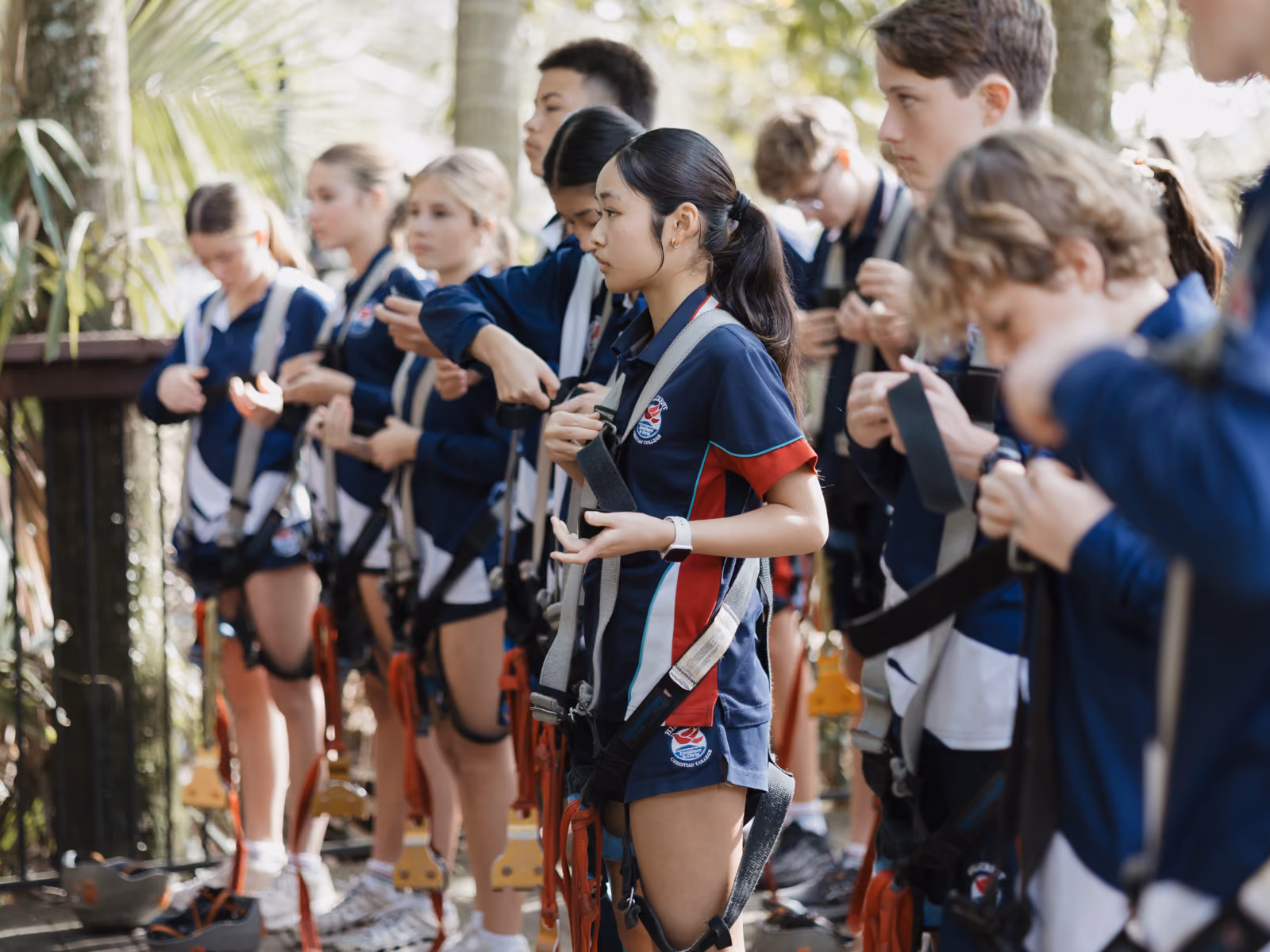 Group of young people in uniforms putting on safety harnesses in an outdoor forest setting.