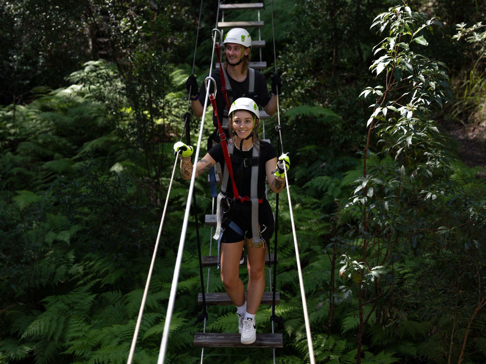 A man and a woman wearing a harnesses crossing a suspension bridge at our high ropes course at Tamborine Mountain