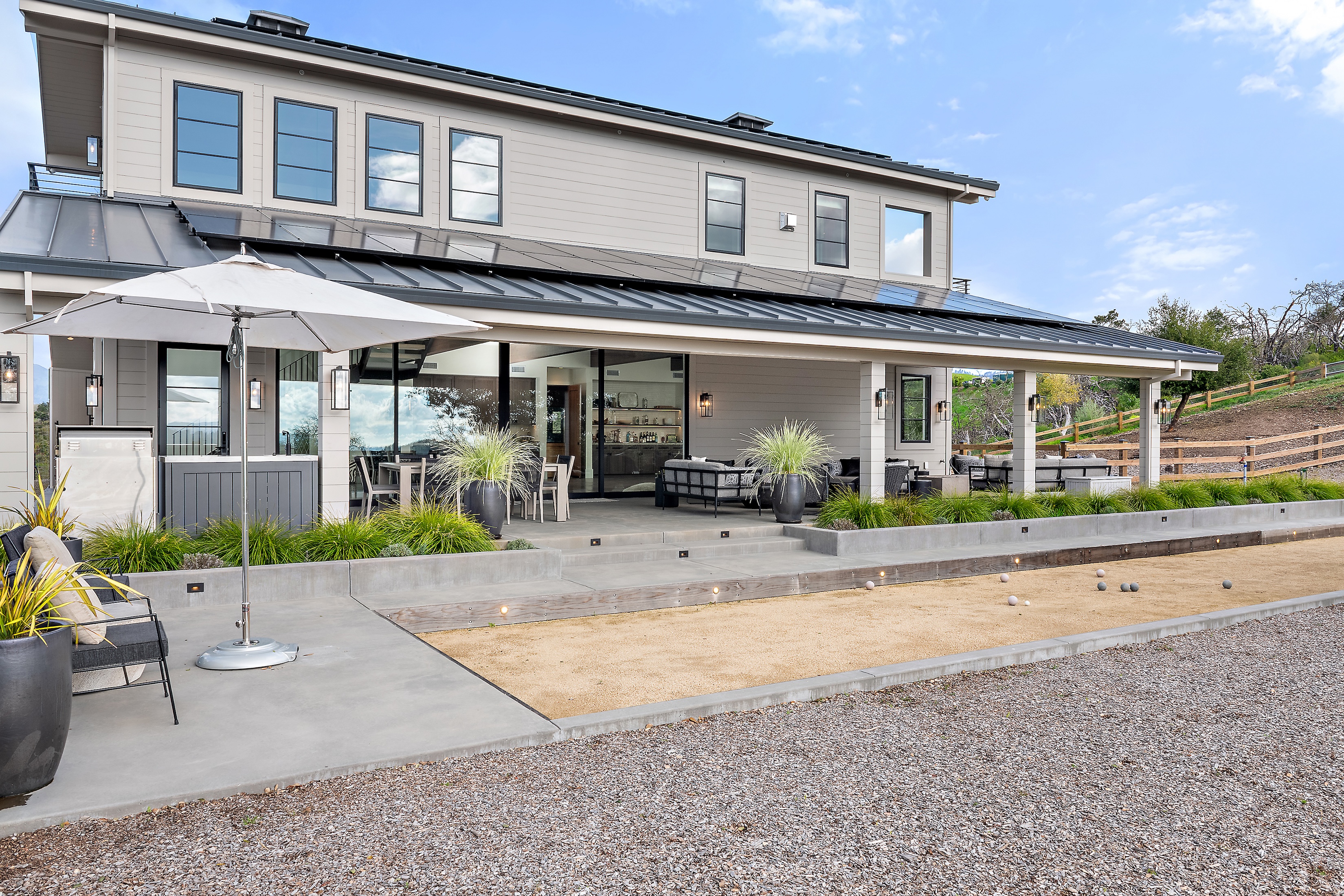 Modern two-story house with large patio, outdoor seating, umbrella, and bocce ball court.