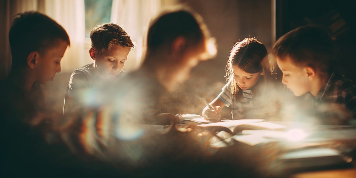 Group of children sitting around a table, focused on writing and reading in a softly lit room.