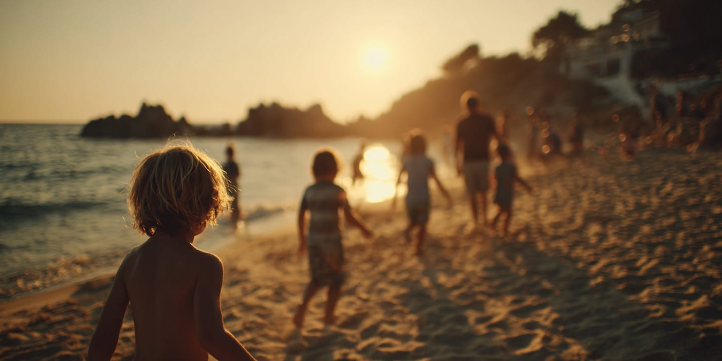 Children and adults walking and playing on a sandy beach at sunset with the sun reflecting on the water.
