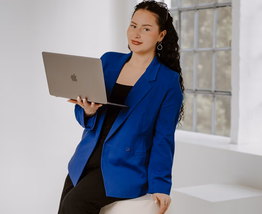 Lisa in a blue blazer holding an open laptop and smiling in a bright room with a large window.