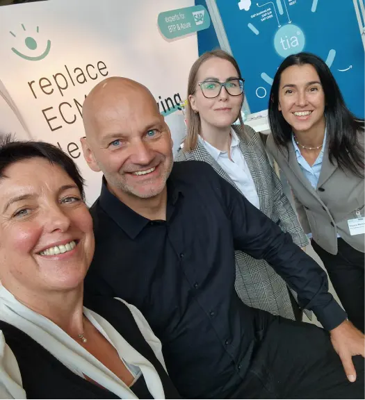 Vier lächelnde Personen machen ein Selfie vor einem Messestand mit dem Schriftzug „replace ECM“.