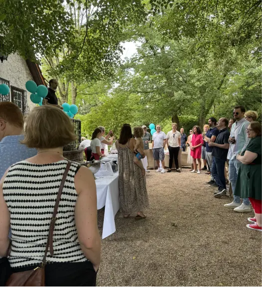 Gruppe von Menschen bei Outdoor-Veranstaltung unter Bäumen mit blauem Luftballons und Tisch mit Getränken.