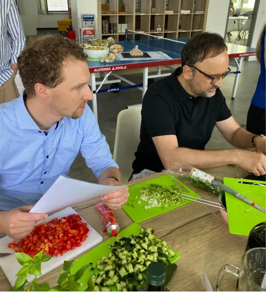 Zwei Männer schneiden Gemüse auf Schneidebrettern in einer modernen Küche mit Tischtennisplatte im Hintergrund.