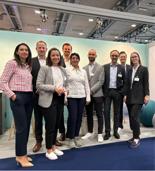 Neun Personen in Business- und Freizeitkleidung posieren lächelnd in einer Messehalle mit modernem Ambiente.