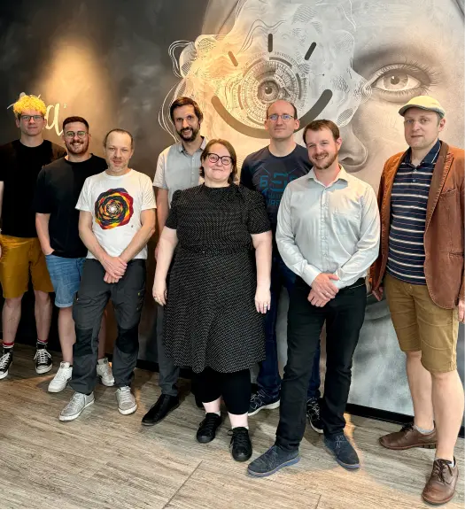 Gruppe von acht Erwachsenen steht vor einer Wand mit künstlerischem Gesicht und abstraktem Design.