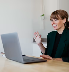 Lächelnde Frau mit grüner Jacke, die vor einem Laptop sitzt und winkt.