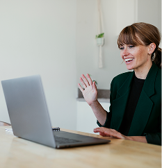 Lächelnde Frau mit grüner Jacke, die vor einem Laptop sitzt und winkt.