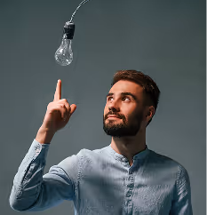 Ein Mann mit Bart und blauem Hemd zeigt mit dem Finger auf eine hängende Glühbirne vor grauem Hintergrund.