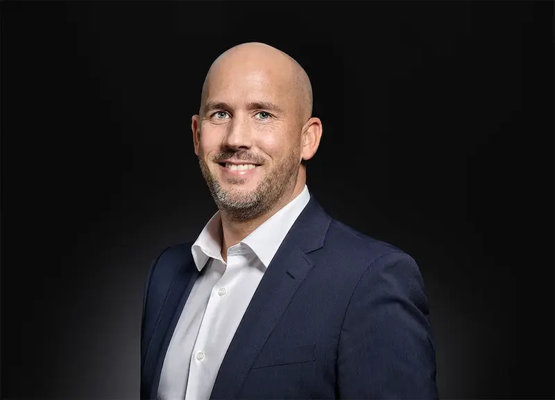 Porträt eines lächelnden, glatzköpfigen Mannes in einem weißen Hemd und dunkelblauem Anzug vor schwarzem Hintergrund.