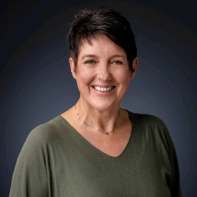 Lächelnde Frau mit kurzem dunklem Haar und grünem Pullover vor dunklem Hintergrund.