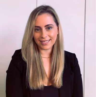 Porträt einer lächelnden Frau mit langen blonden Haaren, die einen schwarzen Blazer trägt, vor hellem Hintergrund.