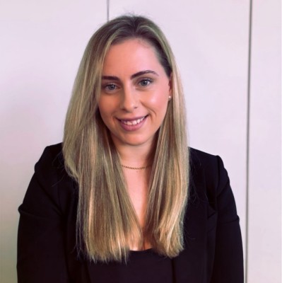 Porträt einer lächelnden Frau mit langen blonden Haaren, die einen schwarzen Blazer trägt, vor hellem Hintergrund.