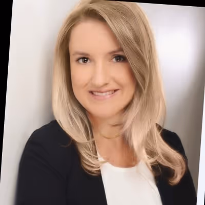 Porträt einer lächelnden blonden Frau mit geraden Haaren, die eine weiße Bluse und ein schwarzes Jackett trägt.