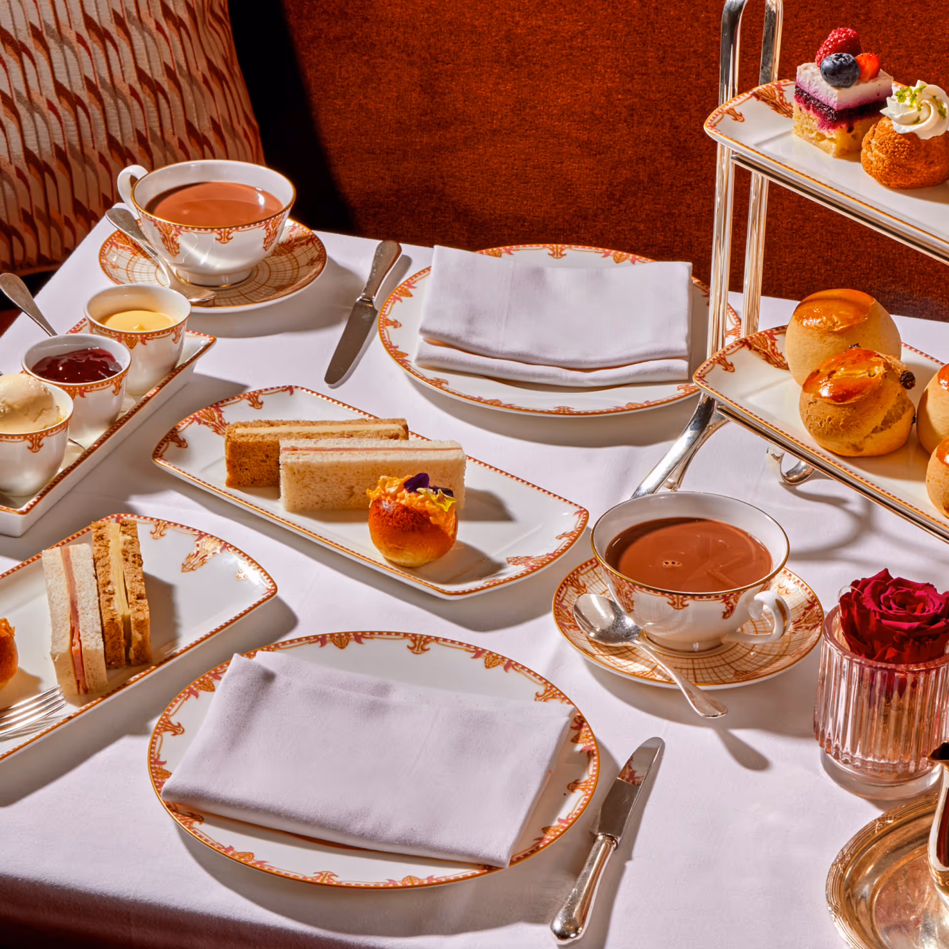 Table set for afternoon tea with patterned cups of tea, finger sandwiches, scones, pastries, and a red rose in a glass vase.