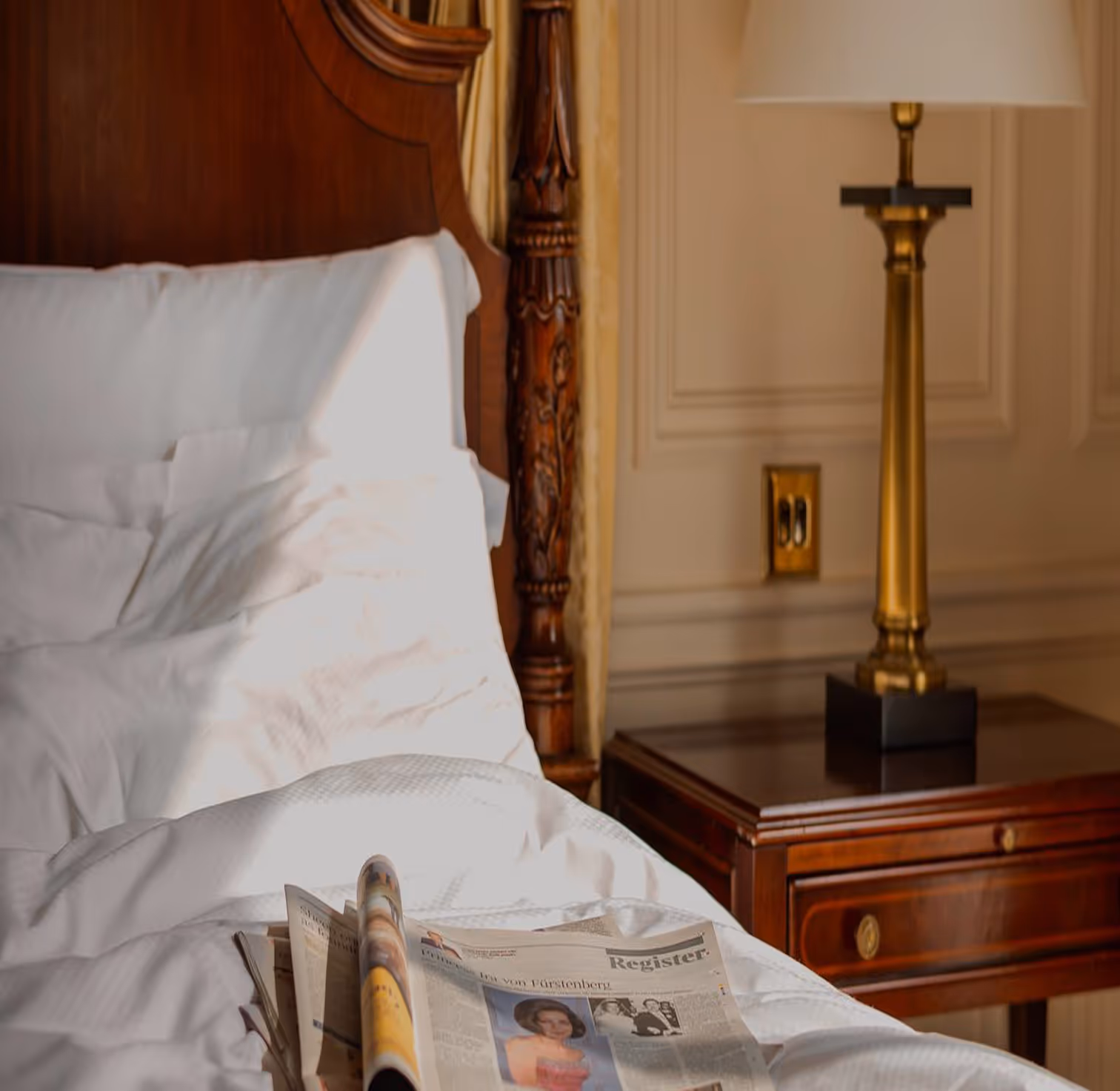 Newspapers placed on a white bed with a wooden headboard and a bedside table with a gold lamp.