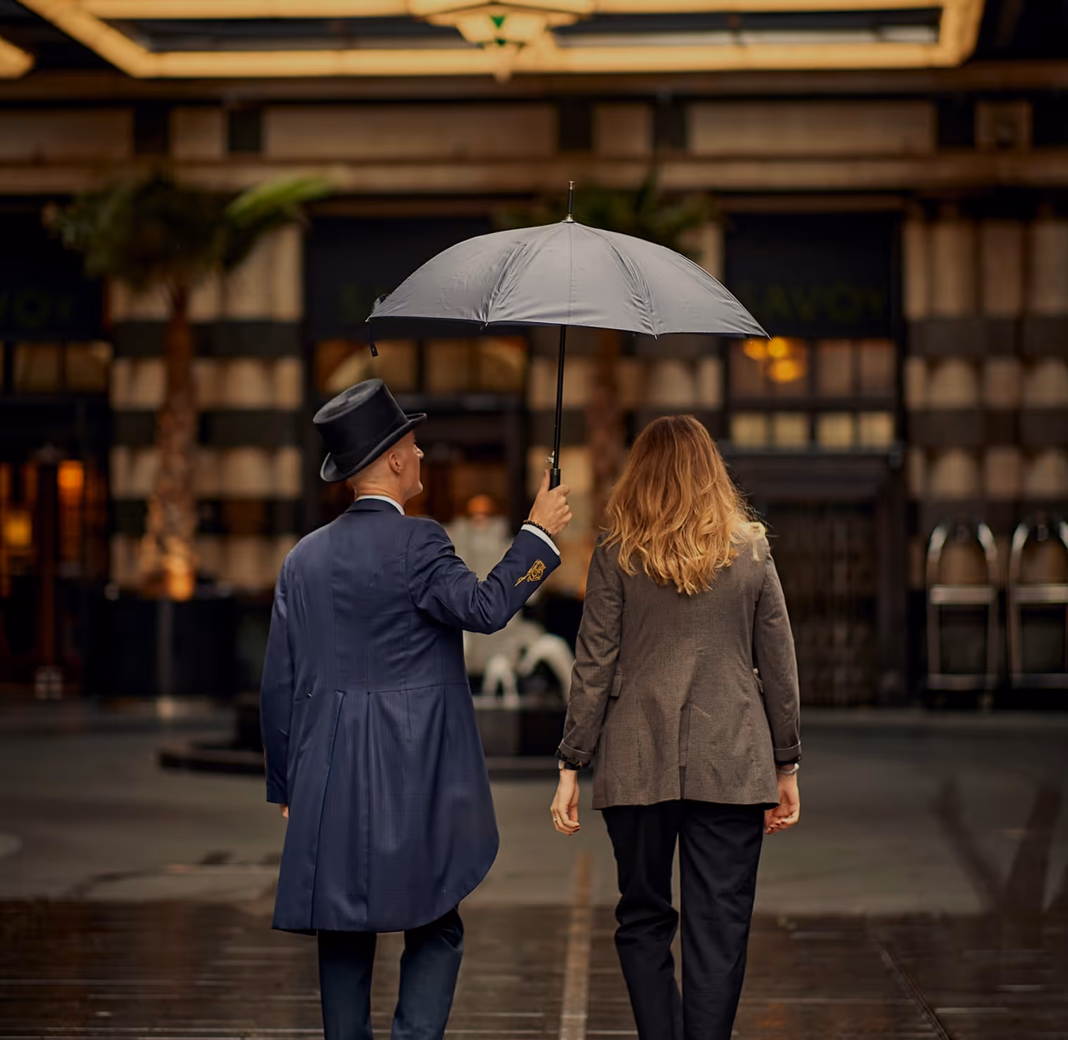 Man in a top hat holding an umbrella over a woman as they walk together on a wet street toward a building entrance.