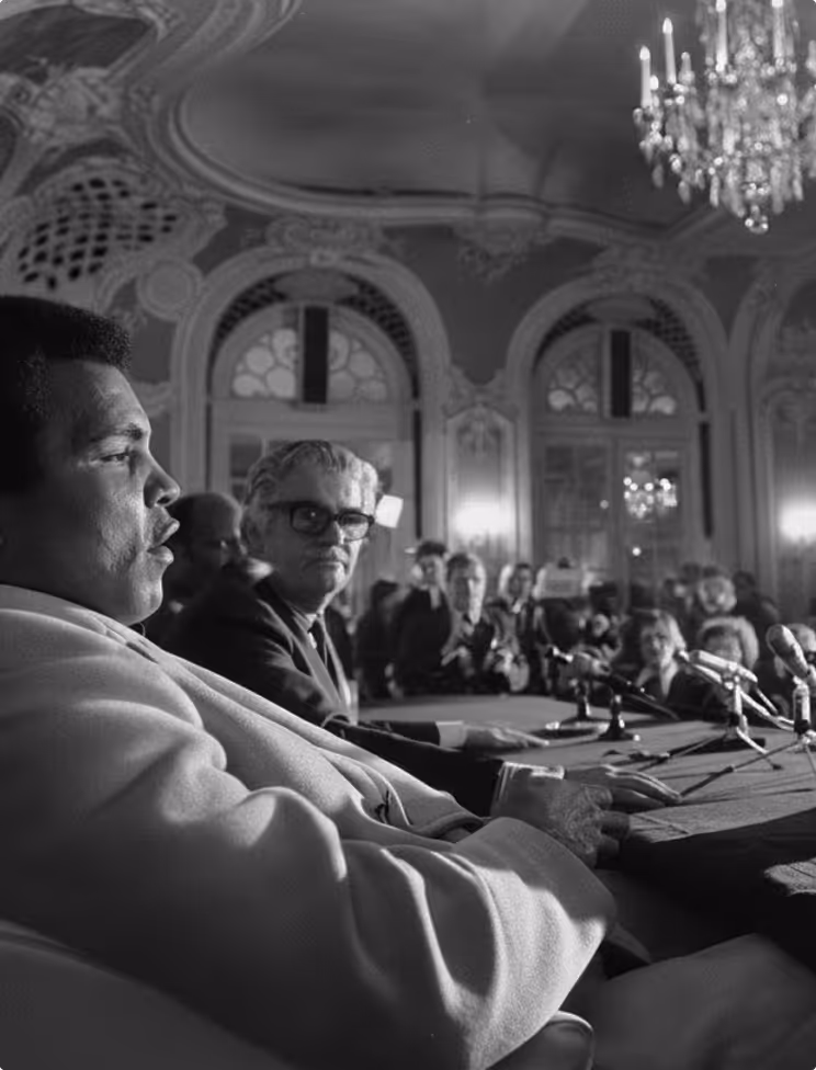 Black and white photo of a man speaking at a conference table with microphones, with ornate windows and chandelier in the background.