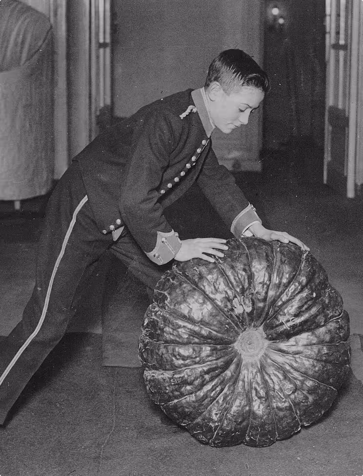Black and white photo of a young man in a uniform bending over to hold a giant pumpkin.