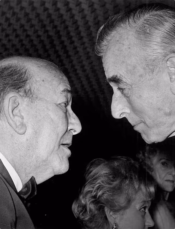 Black and white close-up photo of two elderly men facing each other in conversation, with two women partially visible in the background.