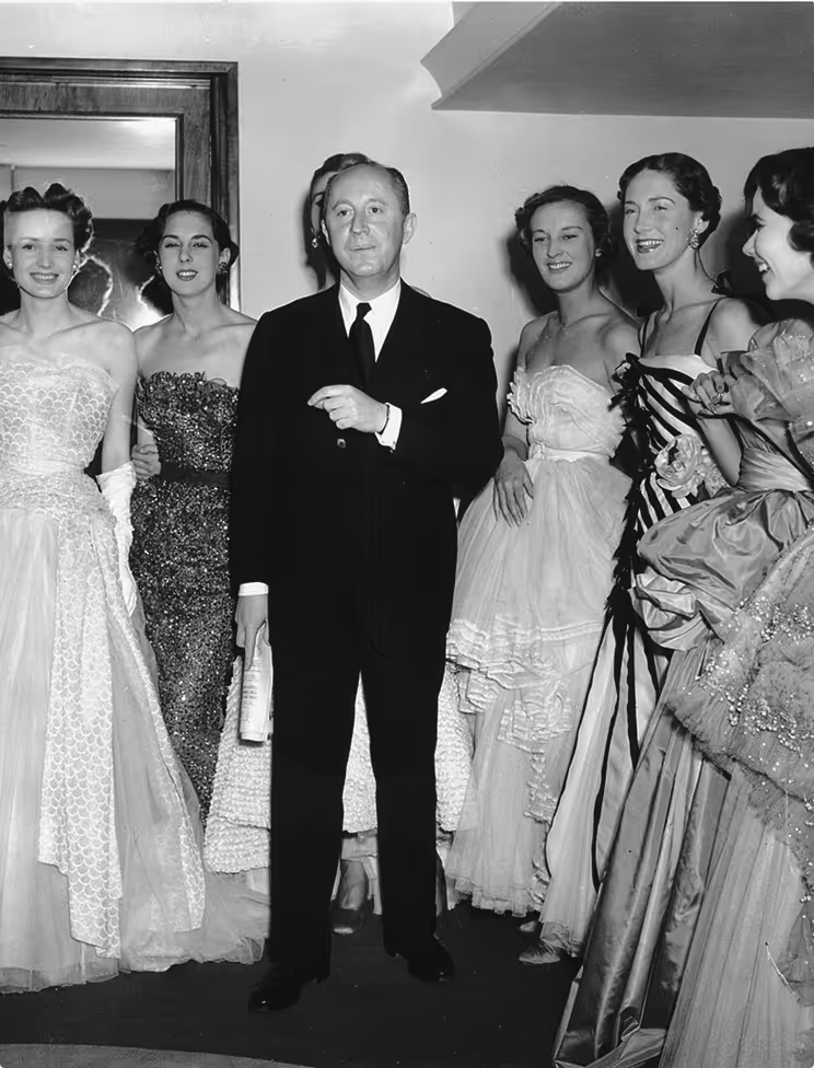 Black and white photo of a man in a suit surrounded by six women in elegant vintage evening gowns.
