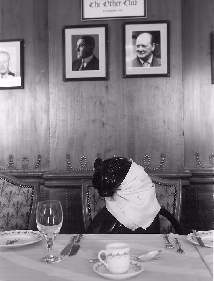 Black dog wearing a napkin around its neck seated at a dining table set with plates, utensils, a glass, and a cup in a formal room with portraits on the wall.