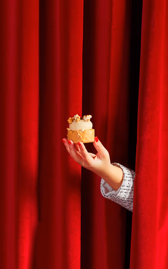 Hand with red-painted nails holding a small dessert topped with popcorn, emerging from between red theater curtains.