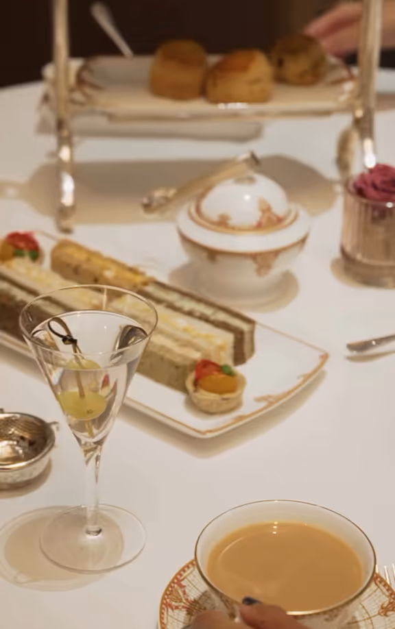 Table set with a teacup of coffee, a martini glass with olives, assorted finger sandwiches, pastries on a tiered tray, and a sugar bowl.