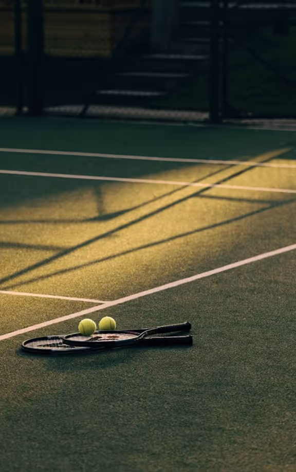 Two tennis rackets and two tennis balls on a green tennis court with sunlight and shadows.