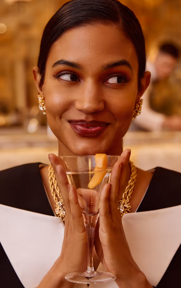 Smiling woman with dark hair holding a cocktail glass garnished with a citrus peel, wearing gold jewelry.