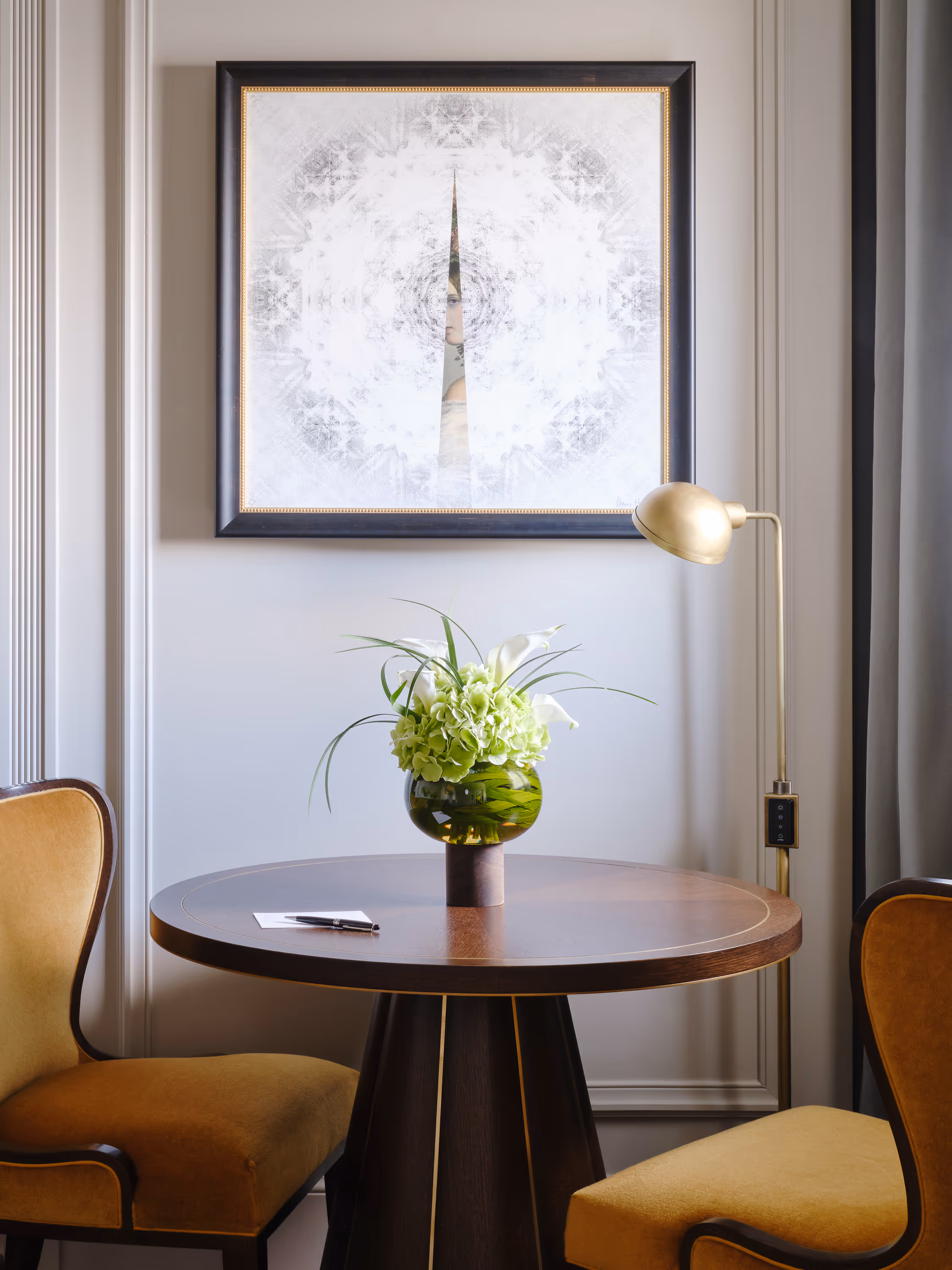 Round wooden table with a green vase of flowers, two mustard-yellow chairs, gold floor lamp, and framed abstract art on the wall.