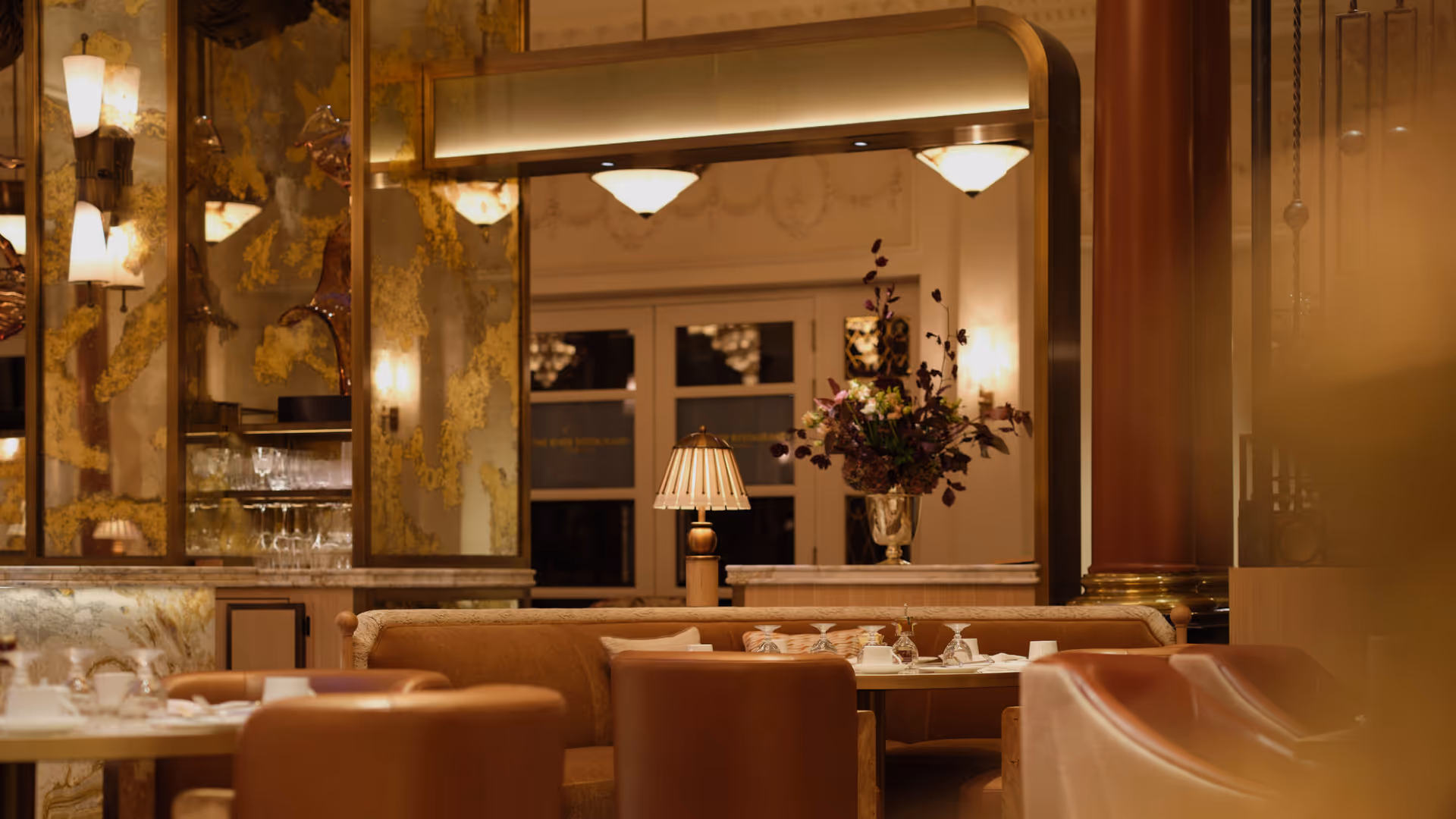 Elegant dining area with brown leather chairs, a beige sofa, a table set with glasses, and a floral arrangement under warm lighting.