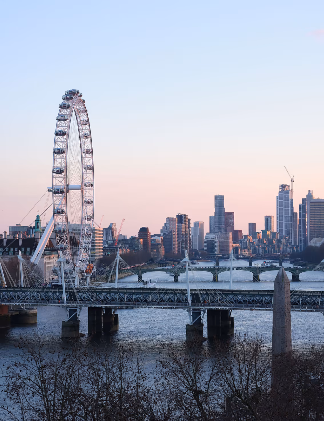 View of the London Eye Ferris wheel and Hungerford Bridge over the River Thames with city buildings in the background at sunset.