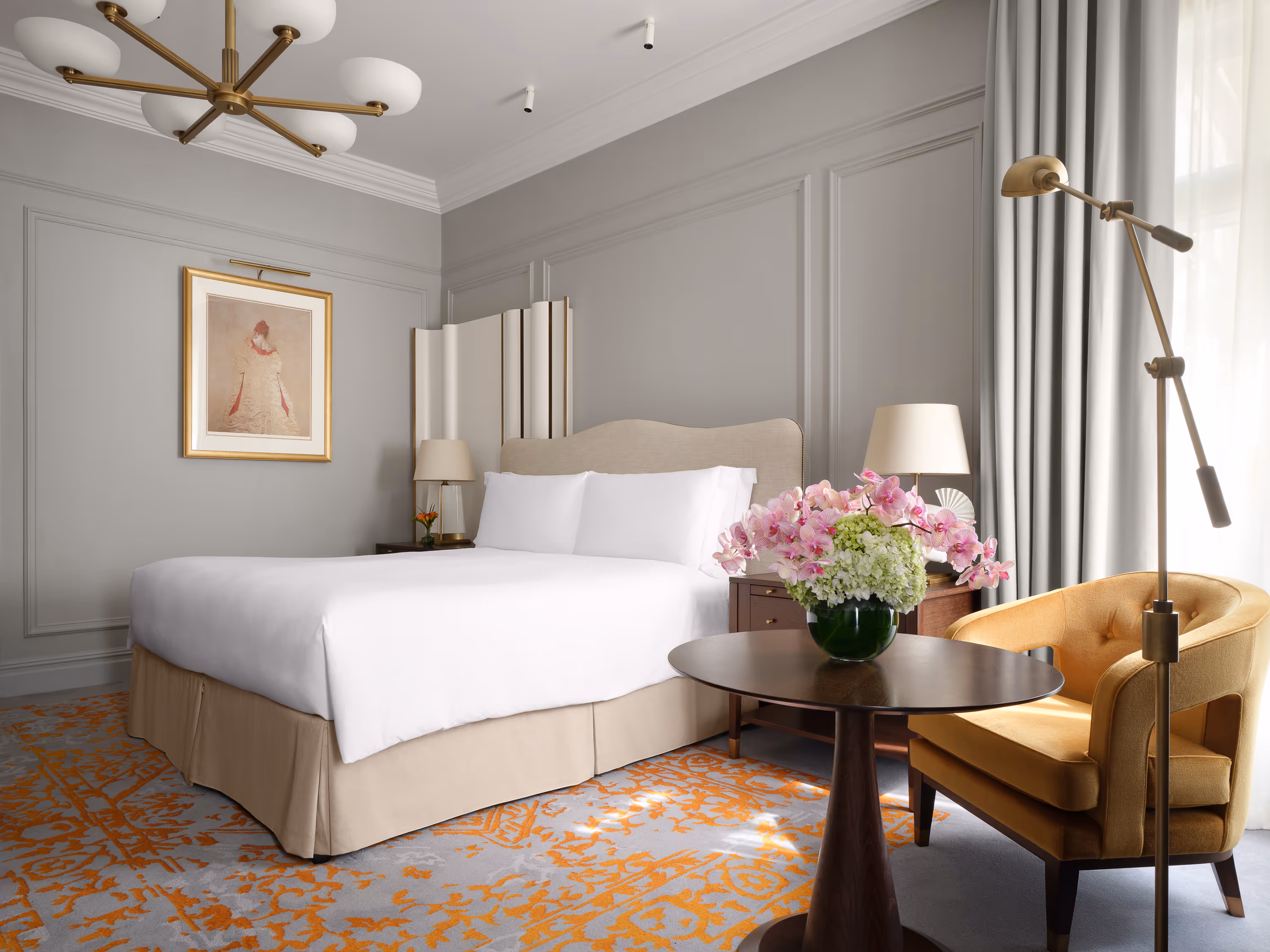 Modern hotel room with a king-size bed, beige upholstered headboard, side tables with lamps, a round wooden table with a flower vase, and a mustard yellow armchair.