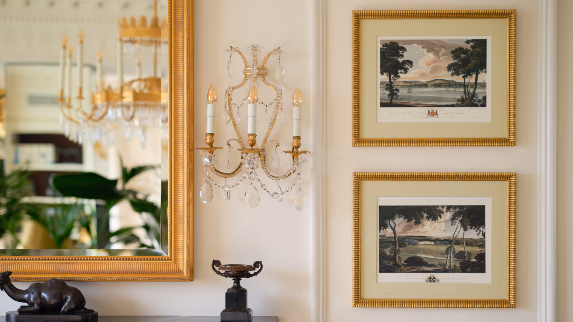 Wall with two framed landscape paintings, a crystal and gold wall sconce, a large gold-framed mirror, and decorative objects on a table.