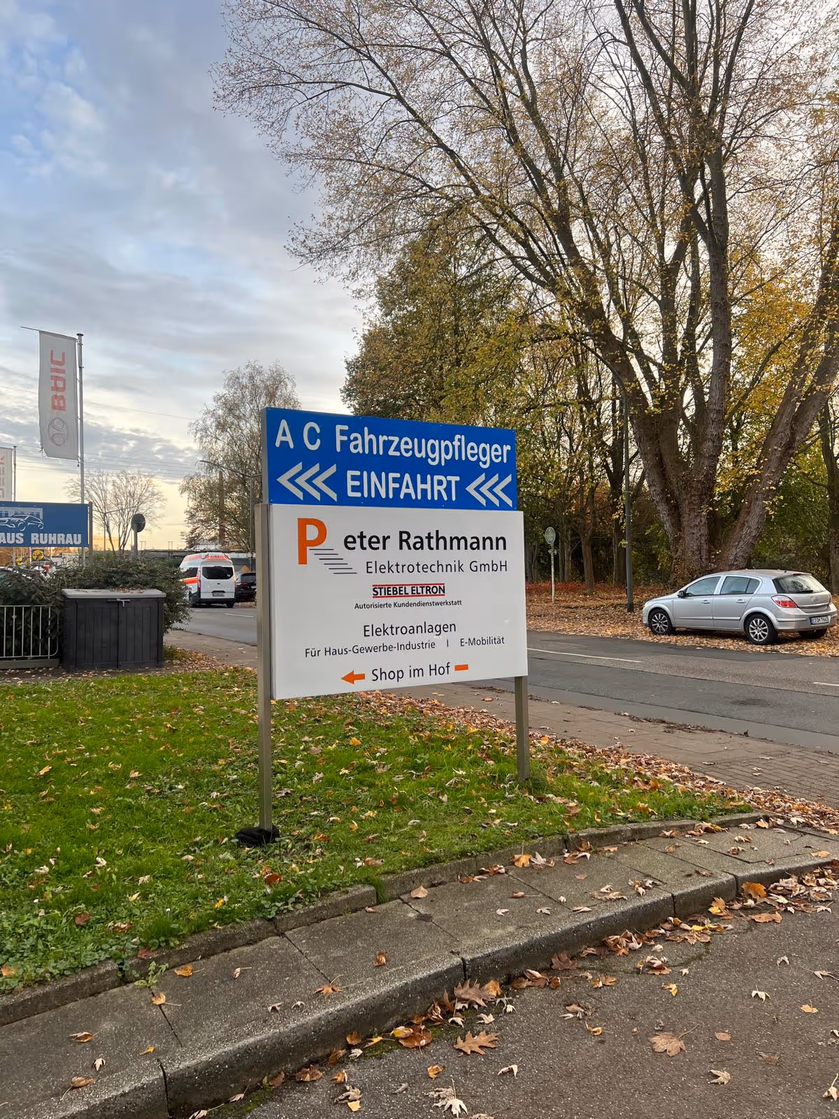 Straßenschild mit Hinweisen zu Fahrzeugpflege, Elektroanlagen und einem Hofshop, umgeben von Bäumen und parkenden Autos.