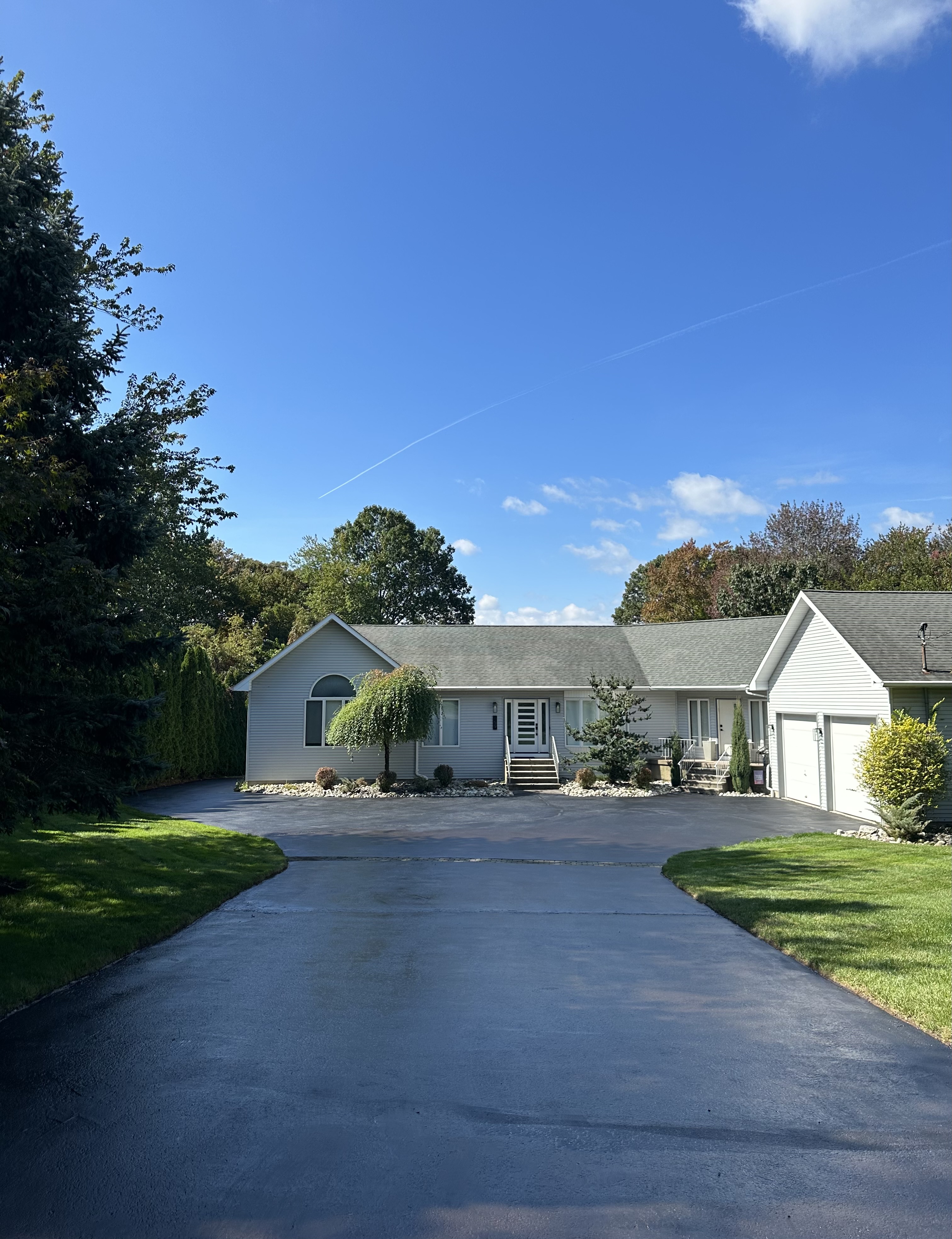 Single-story gray house with a wide driveway, green lawn, and trees under a clear blue sky.