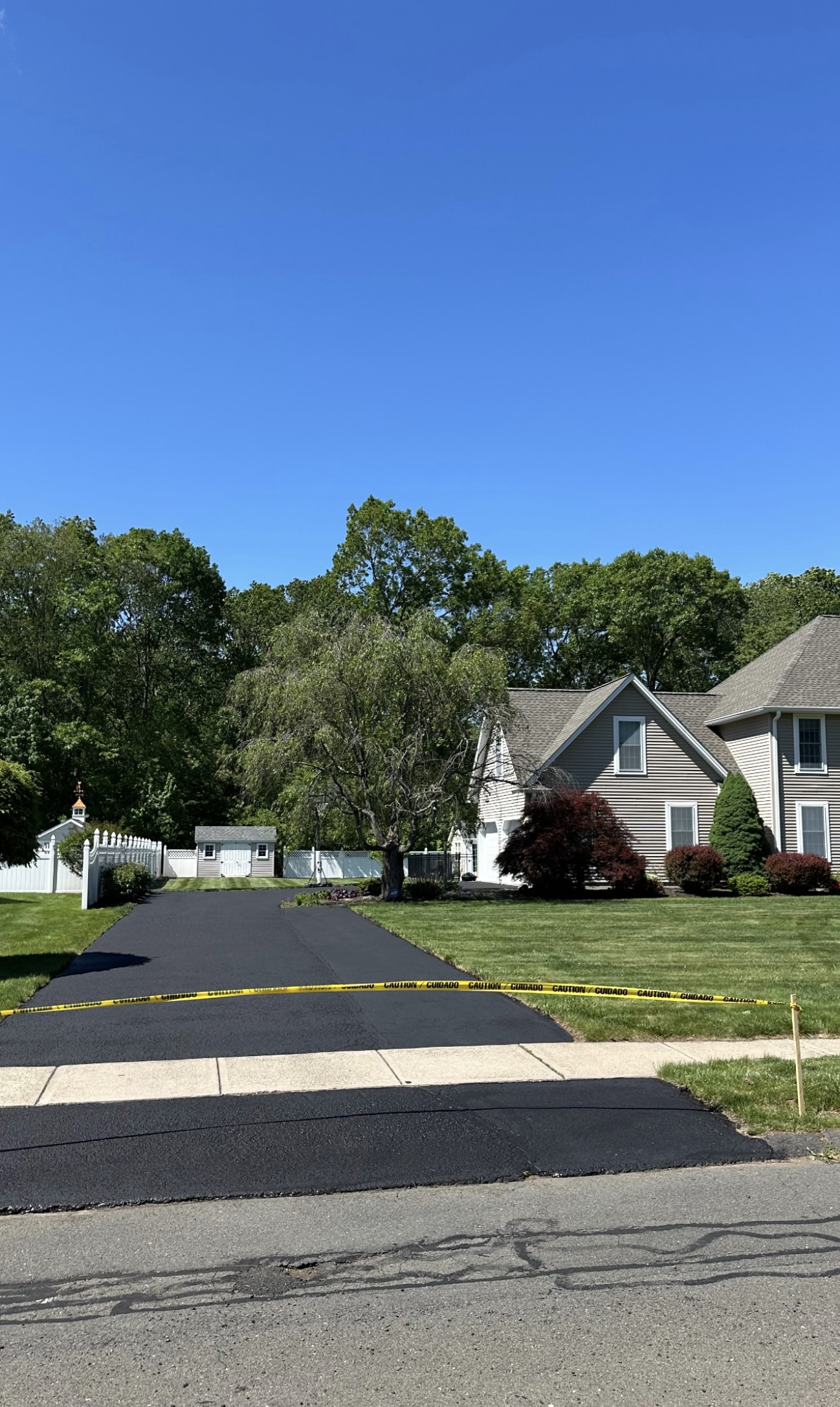 Freshly paved black asphalt driveway with yellow caution tape across the entrance in front of a beige house with green lawn and trees.