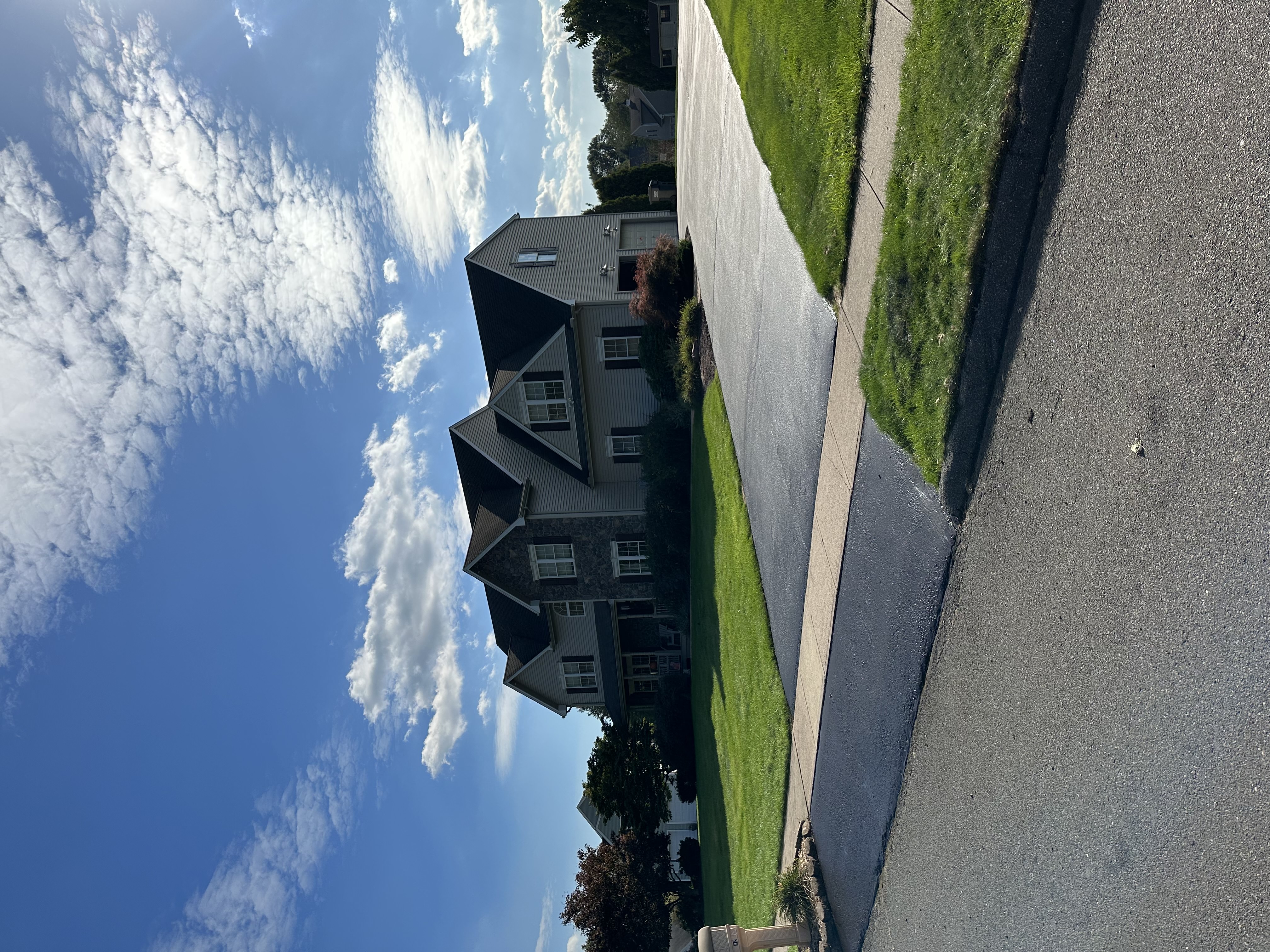 Two-story suburban house with stone and siding exterior, a freshly sealed driveway, green lawn, and partly cloudy blue sky.