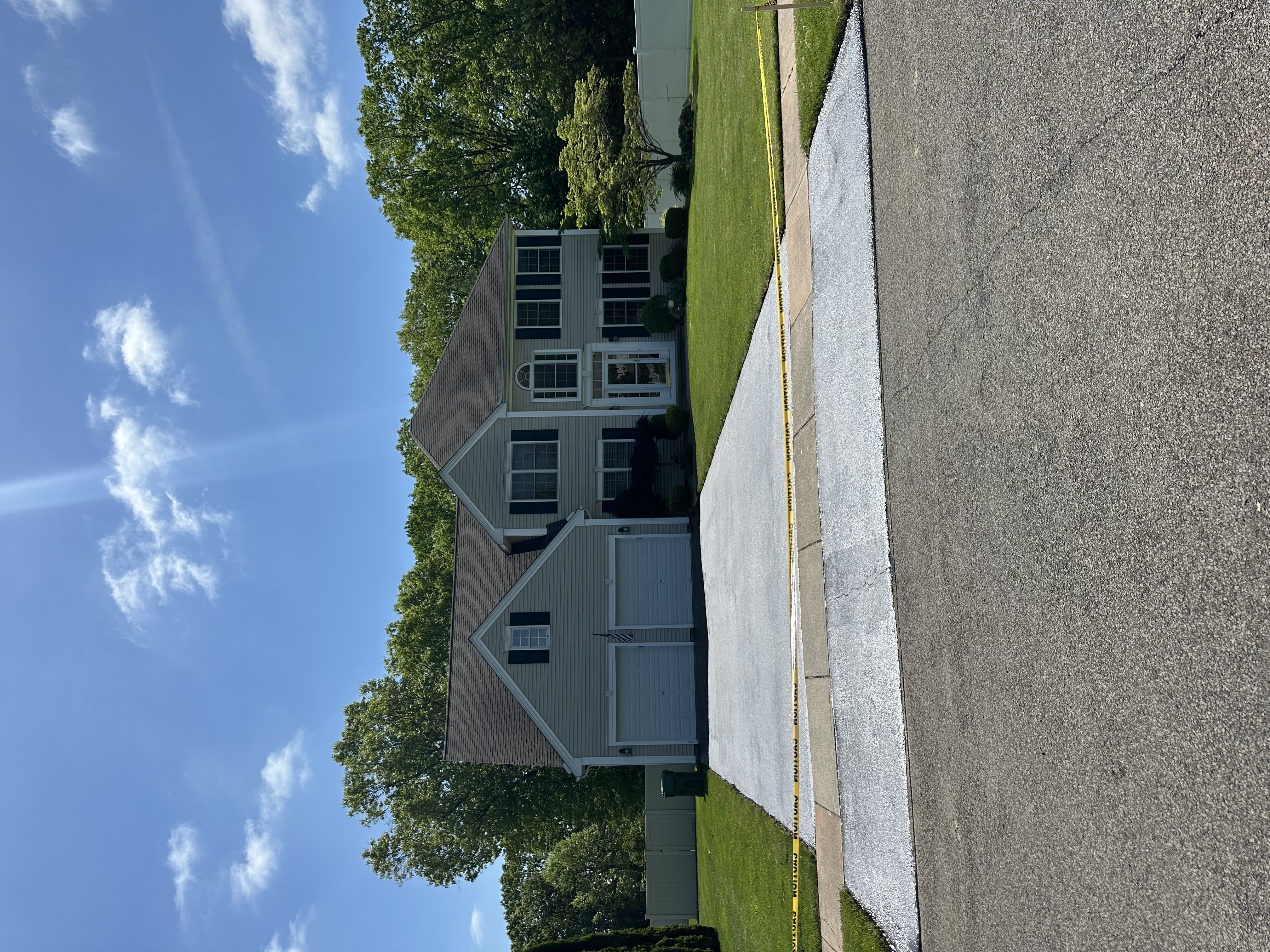 Two-story suburban house with gray siding, black shutters, a double garage, fresh driveway, and a yellow caution tape blocking the entrance.