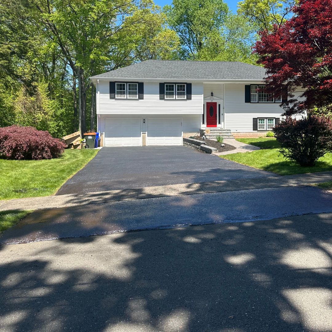 Two-story house with white siding, a red front door, two-car garage, and a freshly paved driveway surrounded by green trees and bushes.