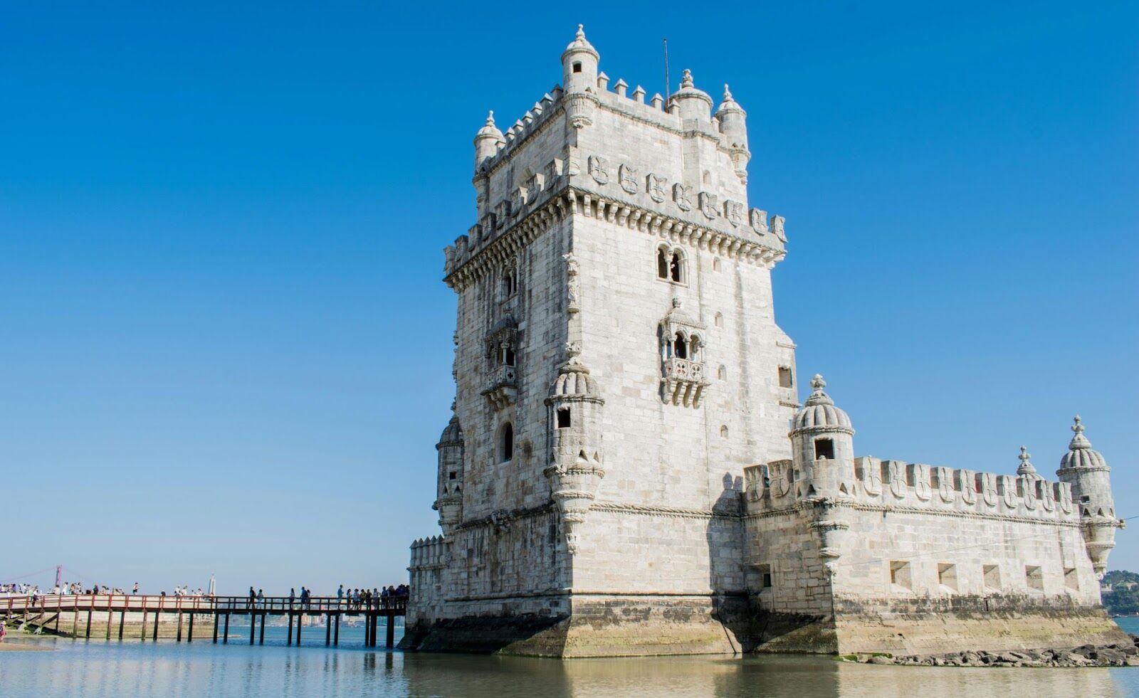 Belem Tower in Lisbon standing by the river with a wooden footbridge and tourists nearby.