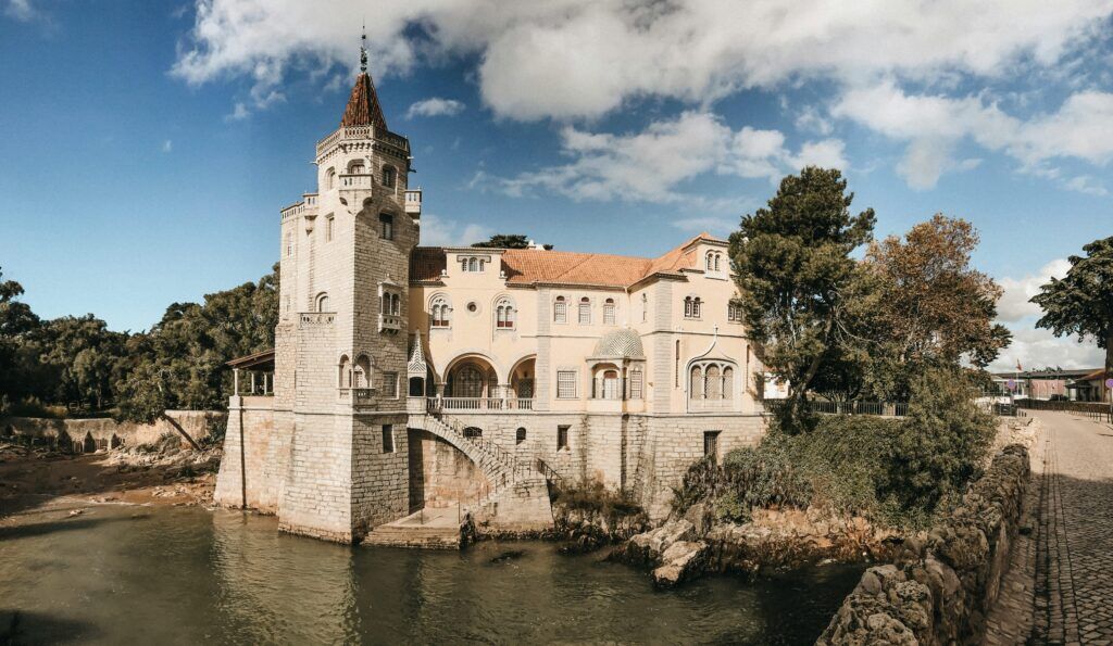 Historic stone mansion with a tower beside a river under a partly cloudy sky.