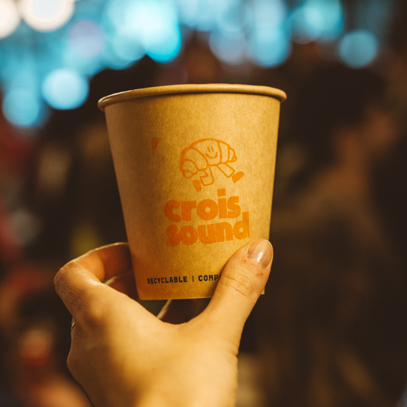 Hand holding a brown recyclable paper coffee cup with an orange logo of a smiling croissant and the words 'crois sound' on it.