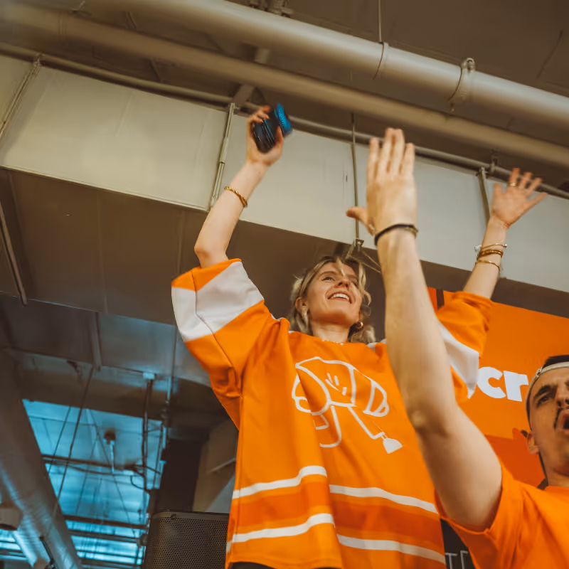 A woman and a man in orange jerseys raising their arms enthusiastically at an indoor event.