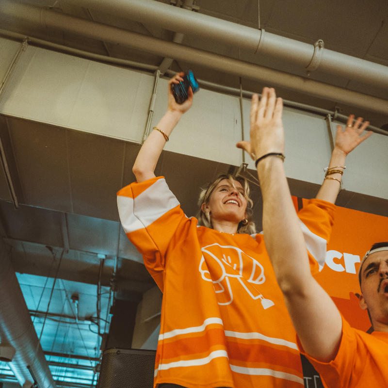A woman and a man in orange jerseys raising their arms enthusiastically at an indoor event.