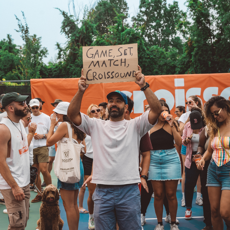 Man holding a cardboard sign that reads 'GAME, SET, MATCH, CROISSOUND' at an outdoor event with people and a dog around.
